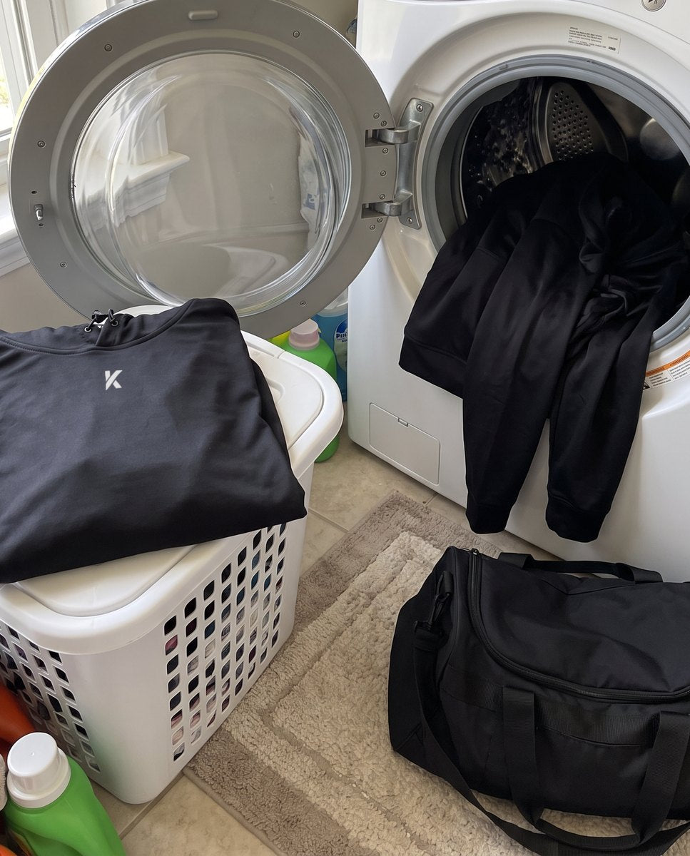 A black hoodie in an open washing machine, a second draped over a laundry basket, and a third folded on a gym bag