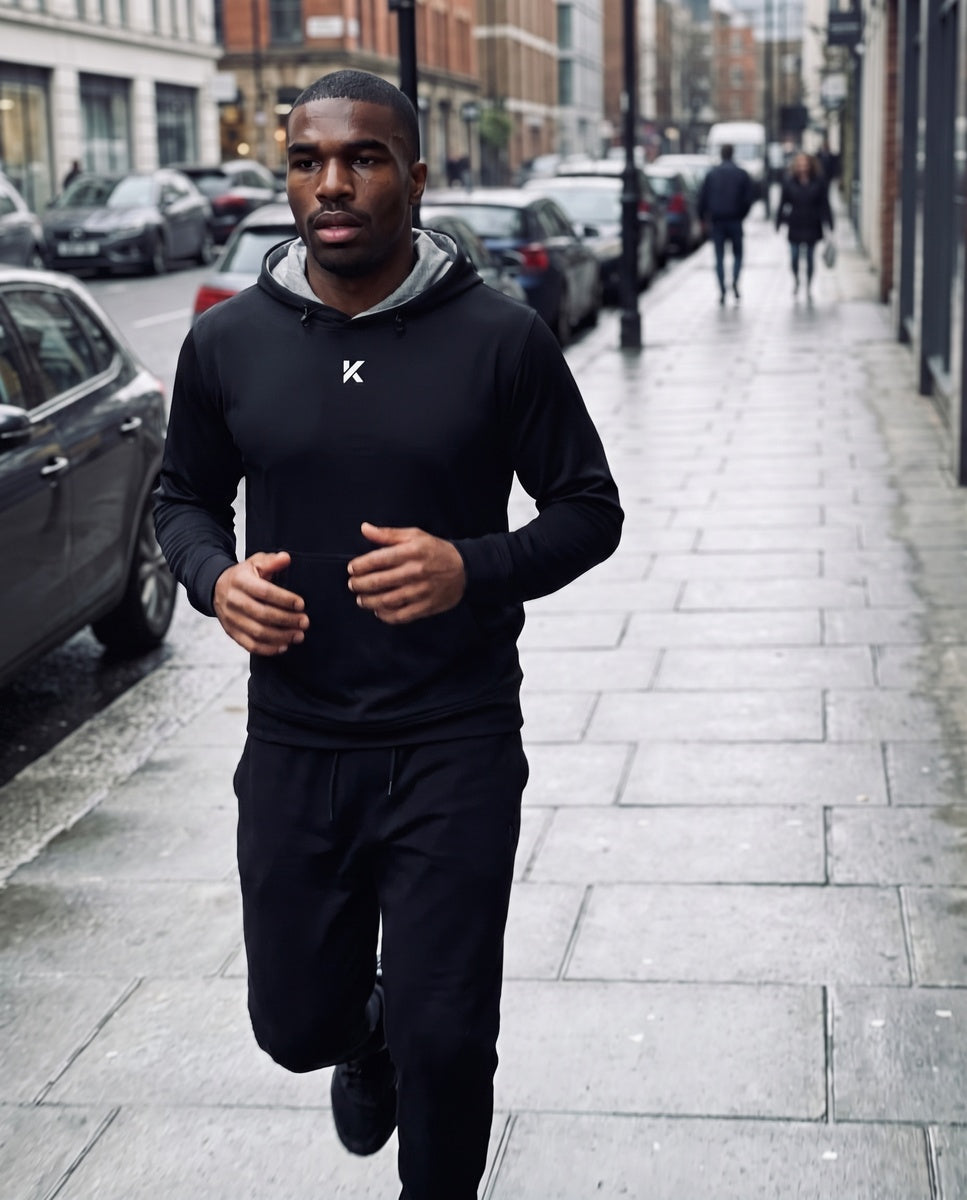 Man running along a wet city sidewalk in the black Pro Sauna Hoodie and black joggers