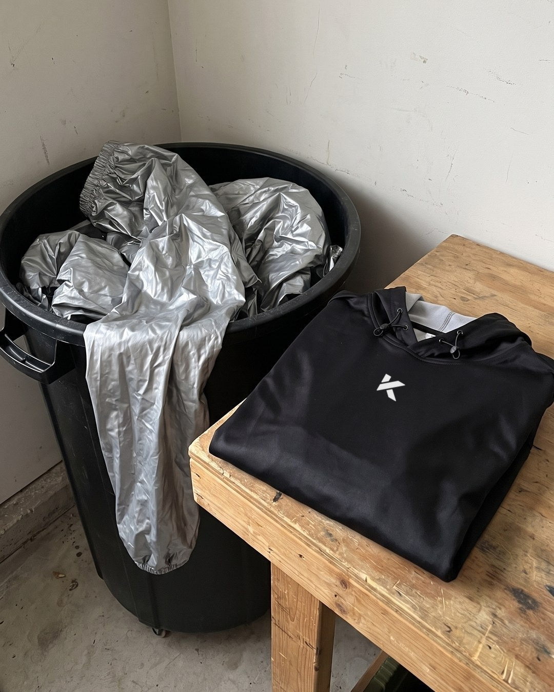 A silver plastic sauna suit stuffed into a black trash bin, with a folded black Kewlioo hoodie on the wooden bench beside it