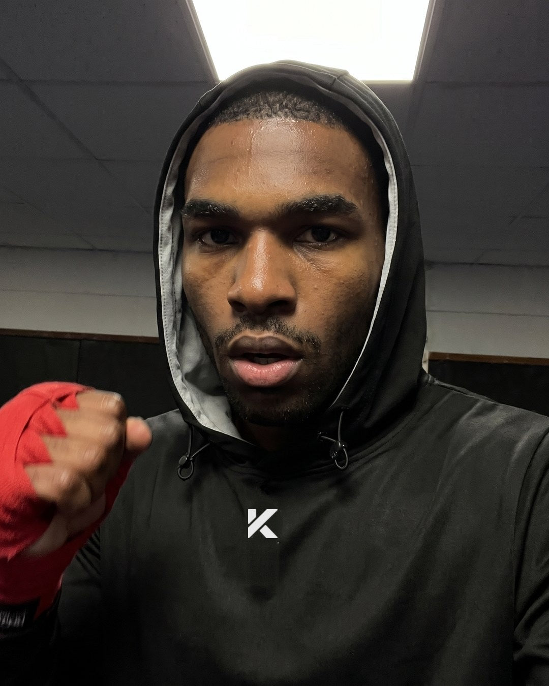 Close-up of a fighter mid-round with the hood of a black Kewlioo hoodie up and red hand wraps on