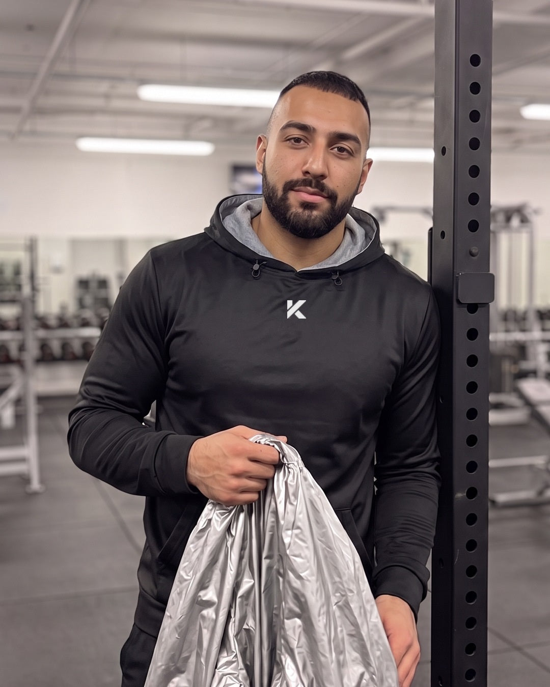 Man in a black Pro Sauna Hoodie at the squat rack, holding a crumpled silver plastic sauna suit