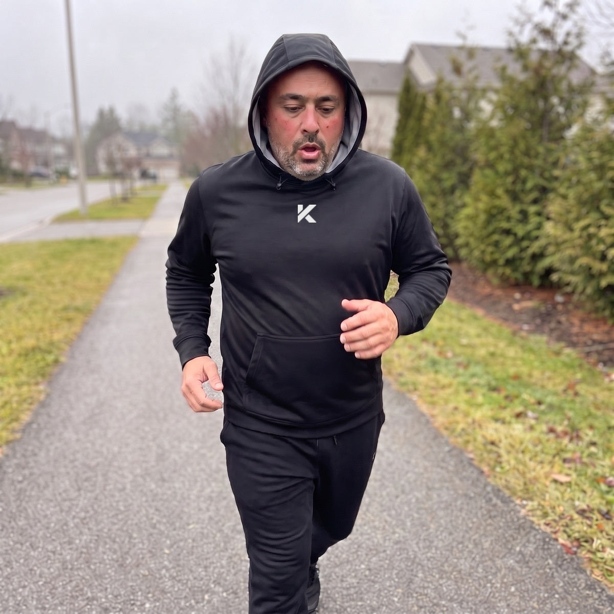 Man jogging along a suburban sidewalk on a cold gray morning with the hood of the black Pro Sauna Hoodie up