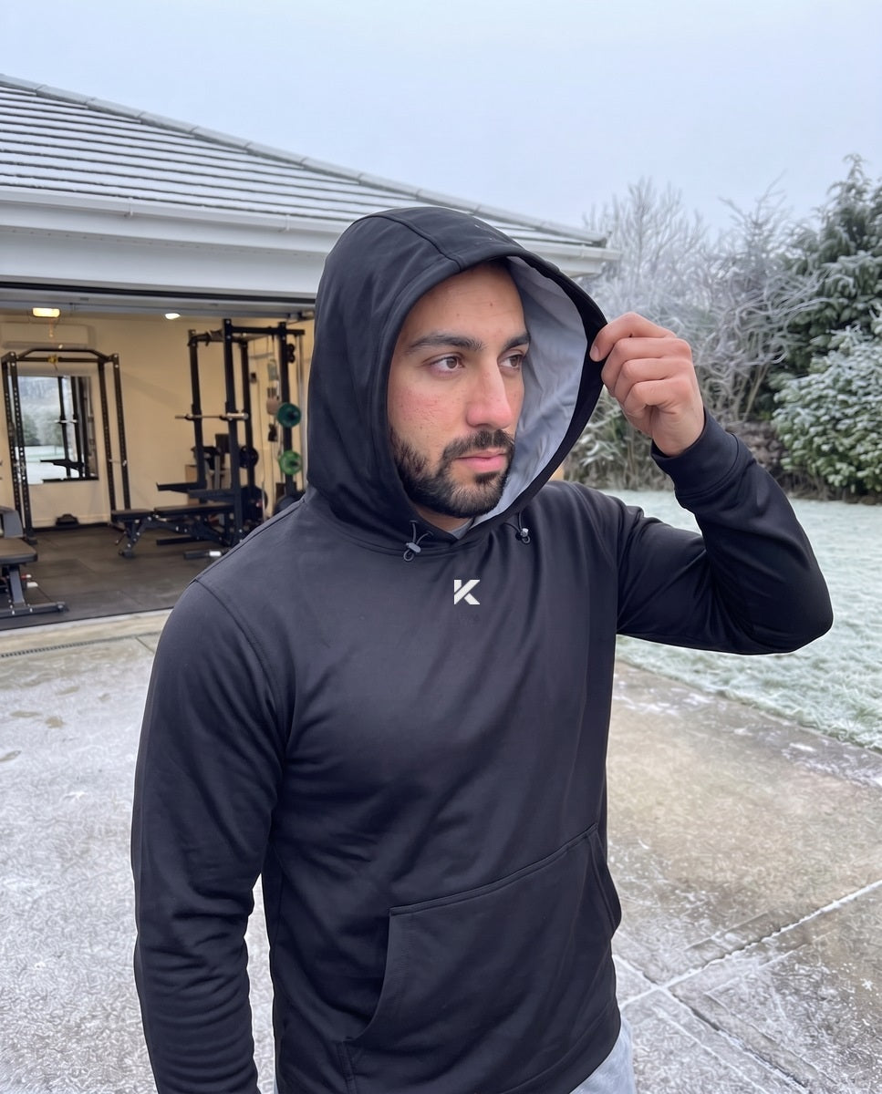 Man with the hood of a black Pro Sauna Hoodie up, standing in front of an open garage gym on a frosty morning, his breath visible