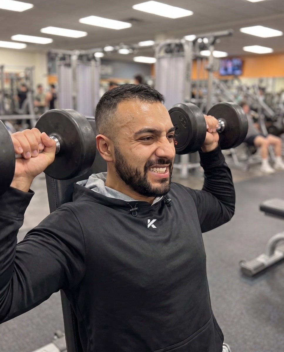 Man in a black Pro Sauna Hoodie pressing two dumbbells overhead on a gym bench