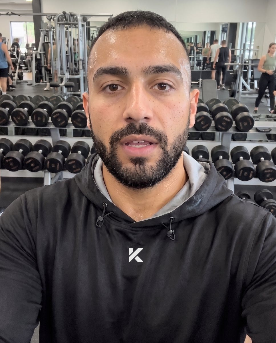 Man in a black Pro Sauna Hoodie filming himself in a gym, dumbbell racks and other members behind him